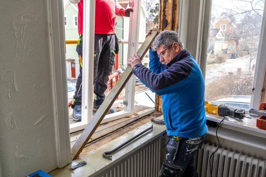 Two men working on a window: one on the outside, wearing red, and one on the inside, wearing blue.